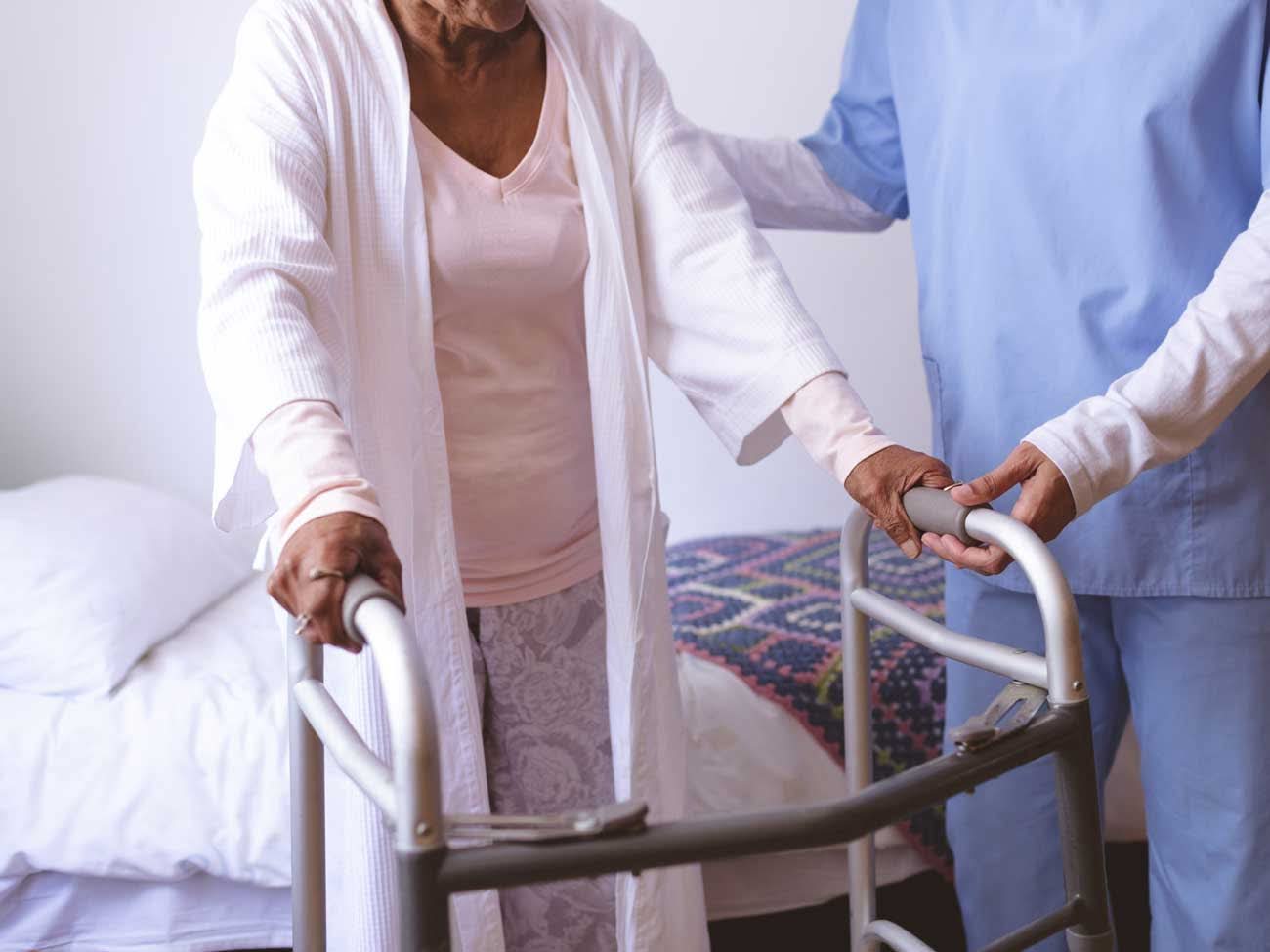 Caregiver assists elderly woman using walker beside bed